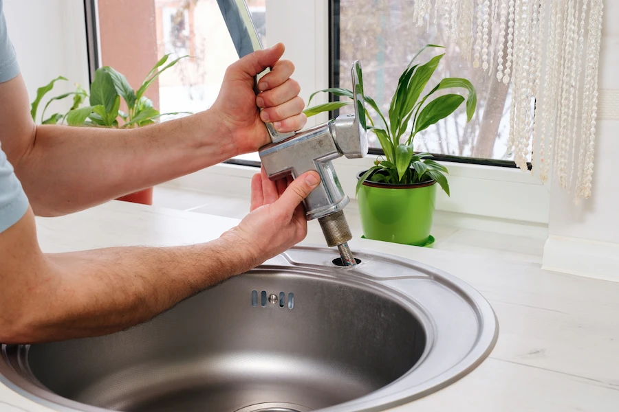 Plumber removing an old kitchen faucet from a stainless steel sink for replacement.